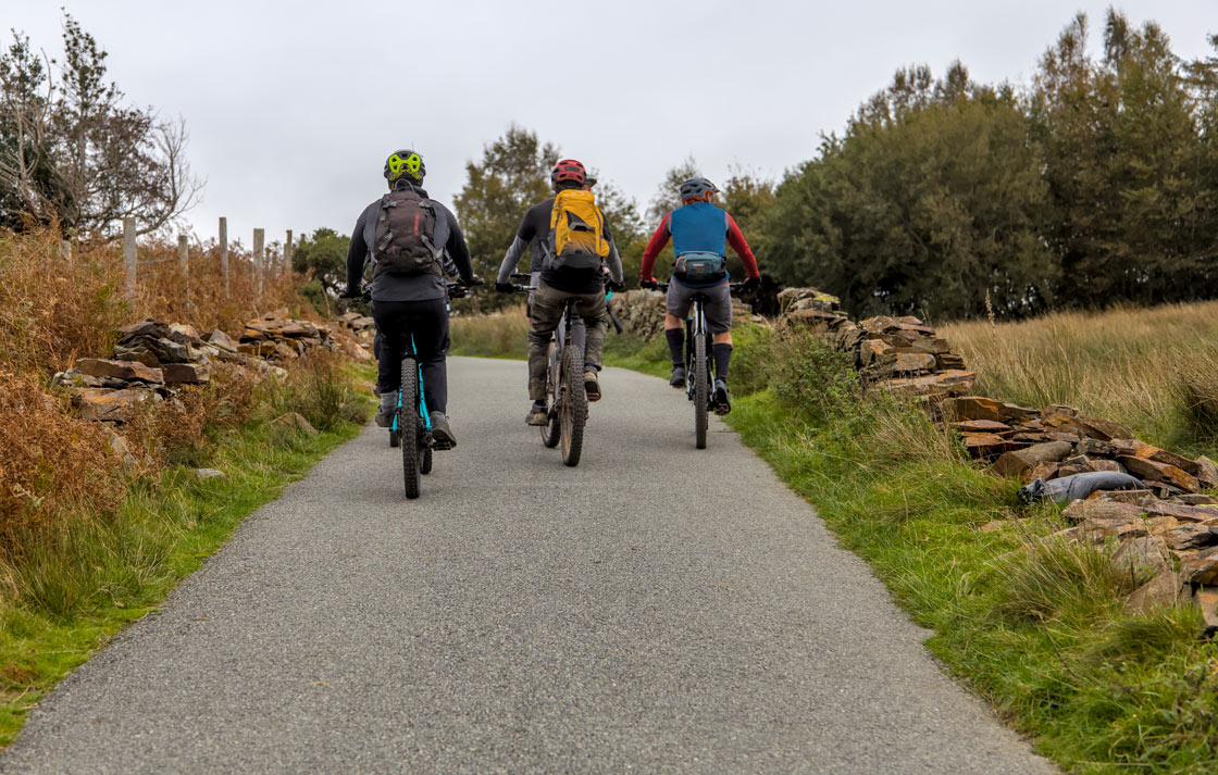 Three adults are seen from behind as they ride their bikes down a paved pathway. To the sides of the path are trees, grass, and shrubbery.