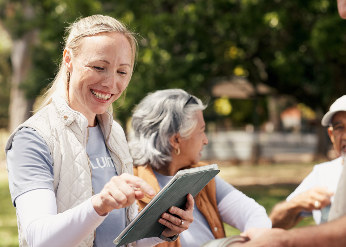 A middle aged woman is smiling wearing casual clothing and holding an ipad. She is surrounded by older women who seem to be enjoying an exercise program outdoors with her as the leader.