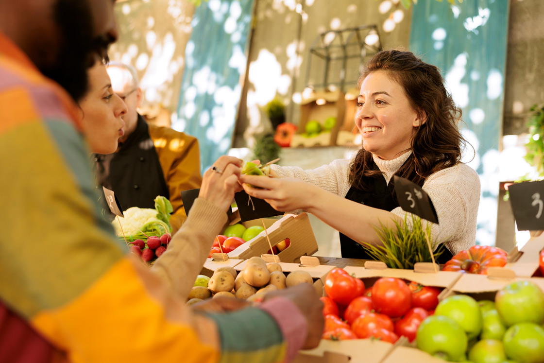 A smiling woman with an apron on is standing behind a stall filled with fresh fruits and vegetables and is handing a fruit to a couple whose arms we can see in the foreground