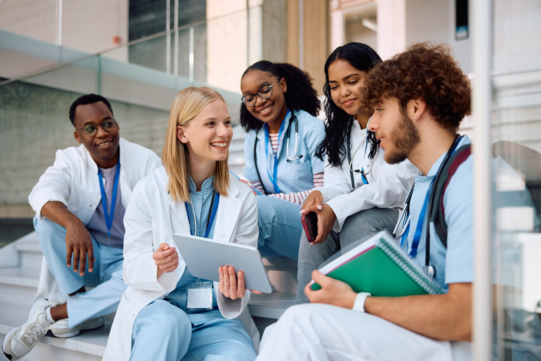 med_students_nurses A smiling woman with an apron on is standing behind a stall filled with fresh fruits and vegetables and is handing a fruit to a couple whose arms we can see in the foreground